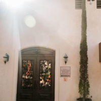 The entrance to a winery's tasting room with decorative wooden doors and a sign indicating it is open daily, with a tree and wine barrel outside.