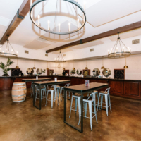 A stylish and elegant winery tasting room with large chandeliers, wooden barrel tables, and a long bar with wine bottles displayed.