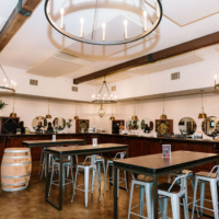 Interior view of a winery tasting room with a bar, high-top tables, large chandeliers, and wine storage areas, with a barrel in the foreground.