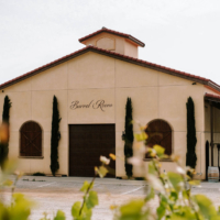 A winery's barrel room with a rustic design, featuring large windows, a wooden door, and surrounded by cypress trees and vineyards.