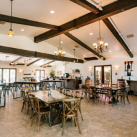 A spacious and beautifully designed winery tasting room with wooden beams, chandeliers, and tables set for guests, offering a cozy and welcoming atmosphere.