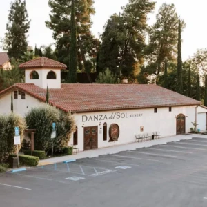 Danza del Sol Winery building with a Spanish-style roof and large windows, surrounded by lush greenery and trees, with a spacious parking area.