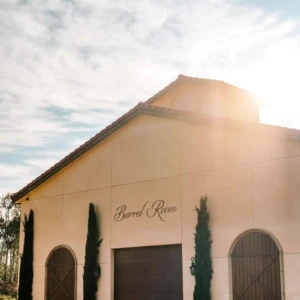 The exterior of a winery's barrel room, with large wooden doors and tall cypress trees, under a clear sky with the sun shining.