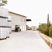 The exterior of a winery storage area with large containers, a forklift, and a container, with a picnic table and tall trees in the background.