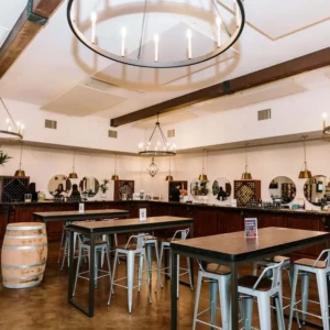 Interior view of a winery tasting room with a bar, high-top tables, large chandeliers, and wine storage areas, with a barrel in the foreground.