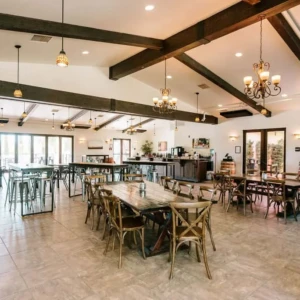 The interior of a winery tasting room with wooden beams, chandeliers, a variety of seating arrangements, and a bar area in the background.